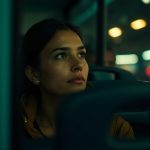 A traveler resting against the window of a dimly-lit overnight bus, illuminated by warm streetlights with city reflections and relaxed posture