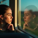 Young passenger gazing thoughtfully at their reflection on a train window, with warm natural light and blurred countryside scenery suggesting the challenges of comfort and space during a long journey.